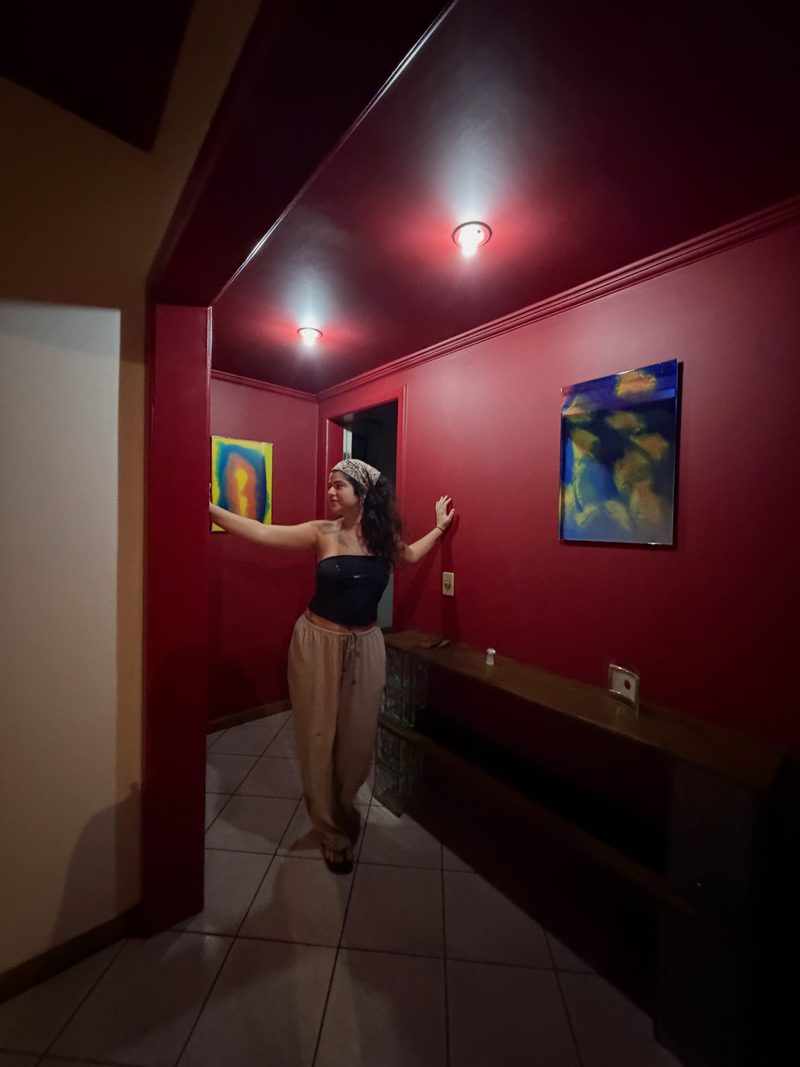 Uma das artistas num corredor de paredes vermelhas, com os braços abertos entre duas obras em espelho e resina penduradas na parede.